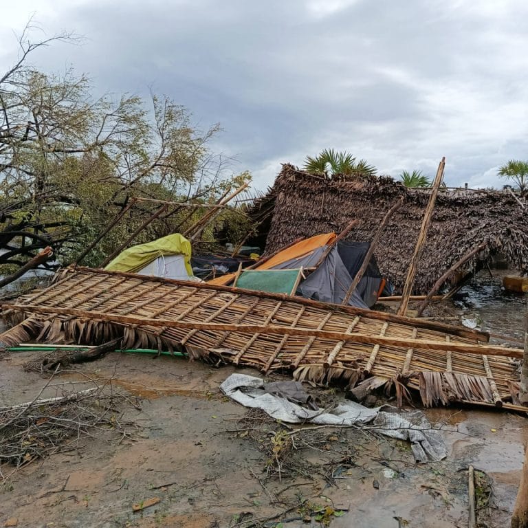 Tents and a palm-thatched shelter lie wrecked after a storm, with leaning wooden frames and muddy debris on the ground.