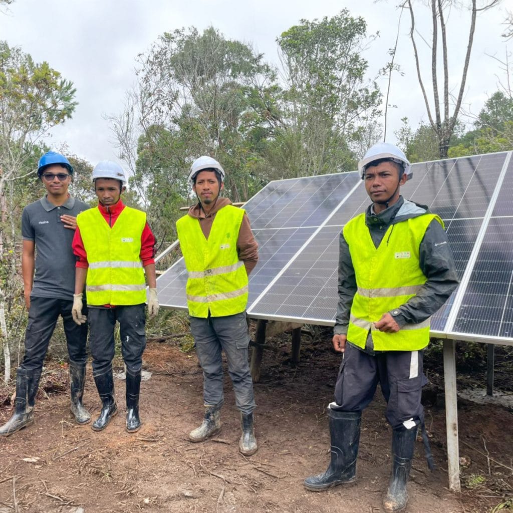 Four workers in hard hats and high-visibility vests stand next to solar panels outdoors at a site of a solar farm.
