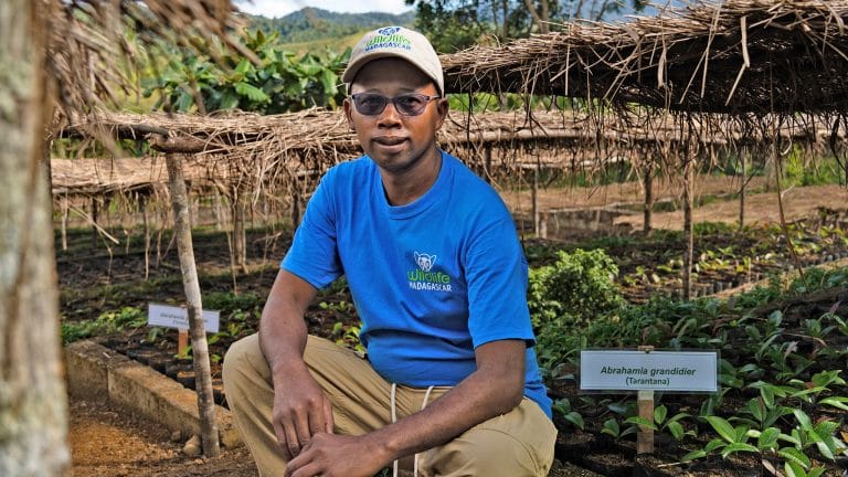 Portrait of a man in a bright blue t-shirt and cap sitting in a garden exhibit, with plant beds and signs in the background (shirt/cap read WillLife Madagascar).