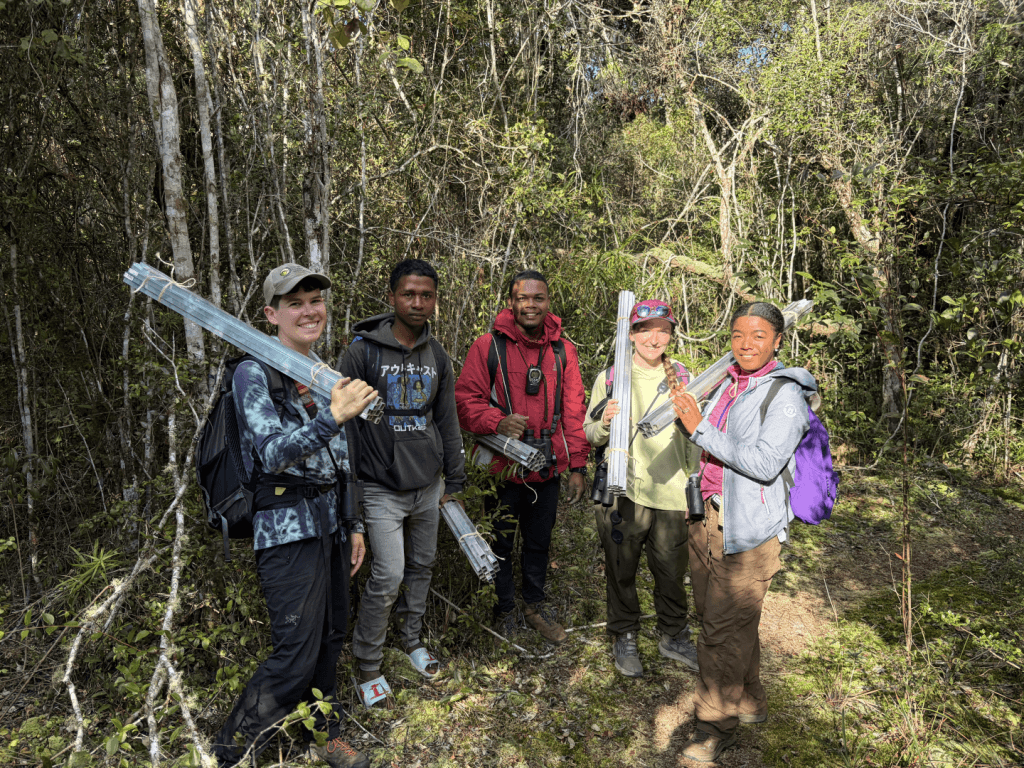 Group of five hikers in a forest, each holding bundles of metal tubing and wearing backpacks while posing for a photo.