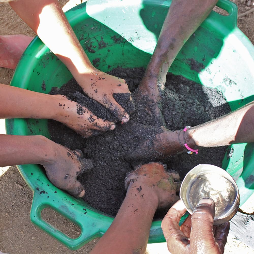 Multiple hands mixing dark soil in a green tub, with a metal cup nearby.