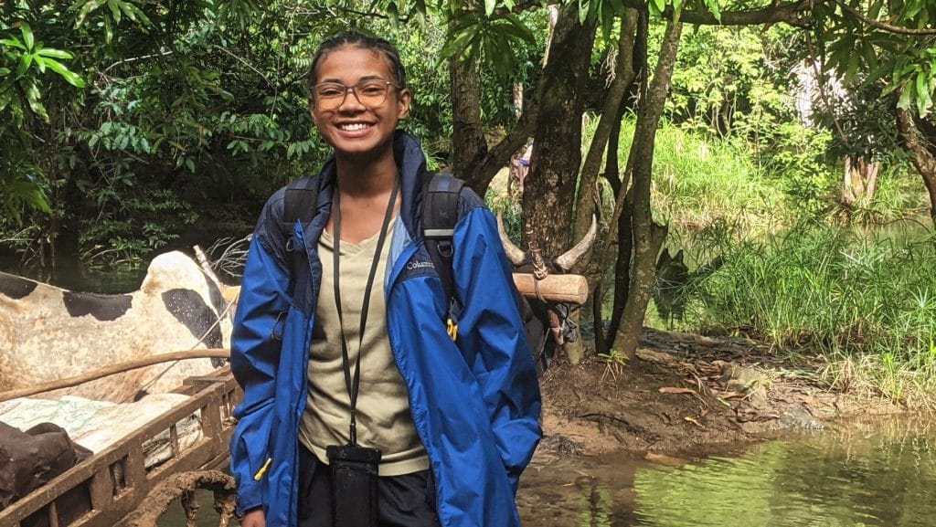 Person standing near a muddy riverbank in a blue rain jacket, smiling at the camera with a backpack and camera bag. The background shows dense green foliage and trees.