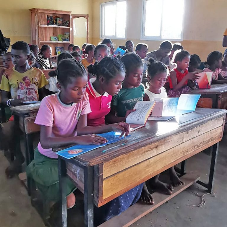 Group of children seated at wooden desks in a bright classroom, reading colorful books together as sunlight comes through the windows.