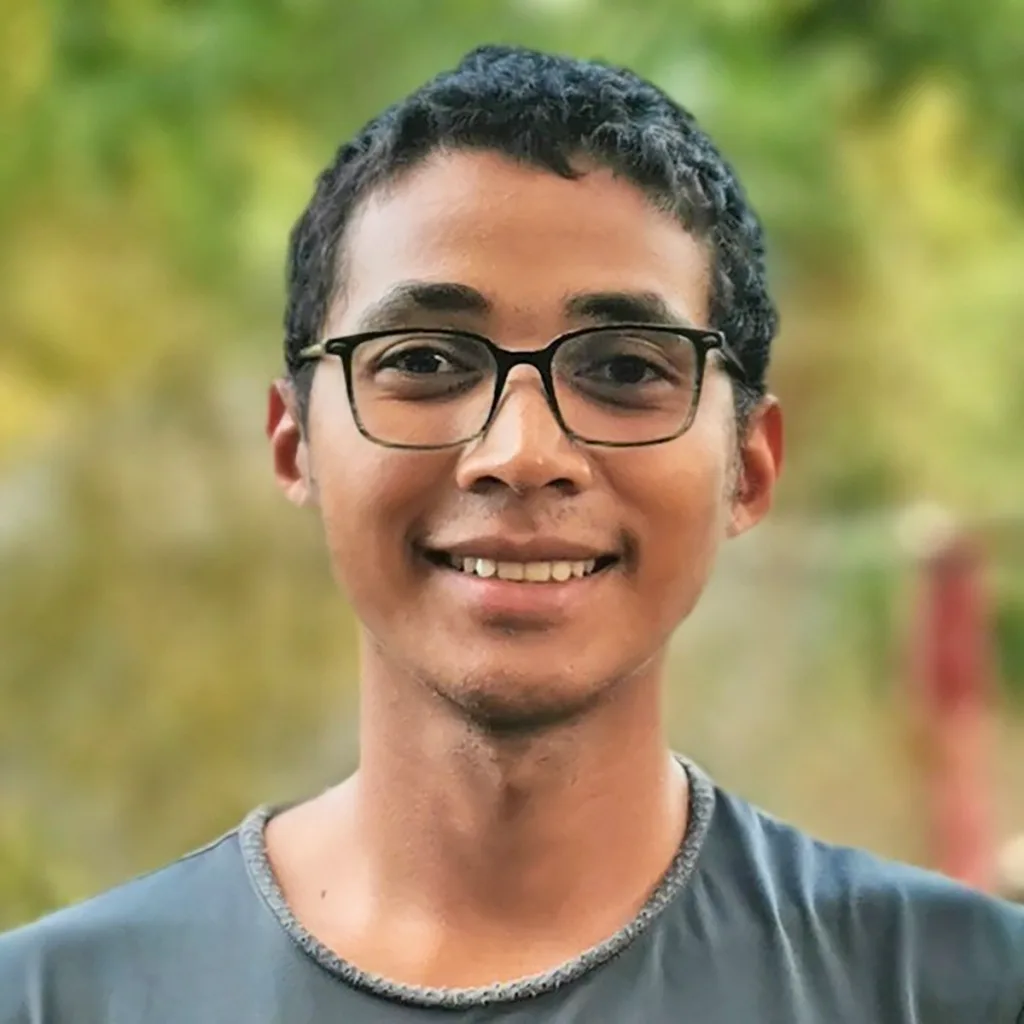 Young man with short black hair and glasses smiling outdoors in a natural setting with greenery behind him, wearing a gray shirt.
