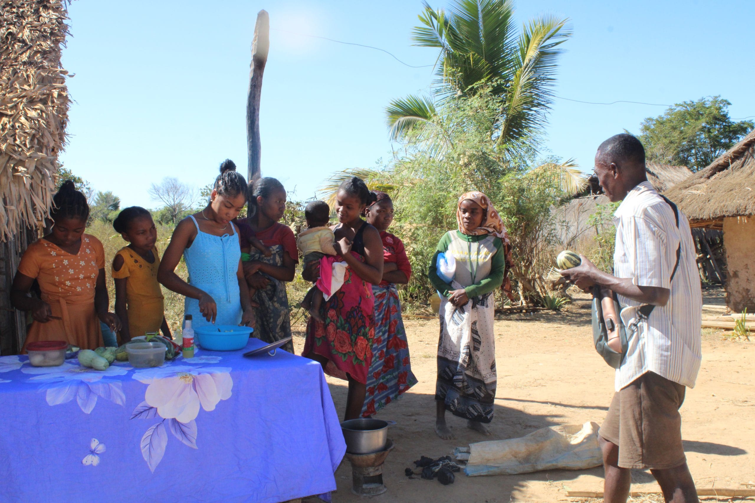Group of people gathered outdoors around a blue table with food containers in a rural setting. A man stands on the right, talking to them under a sunny sky with palm trees.