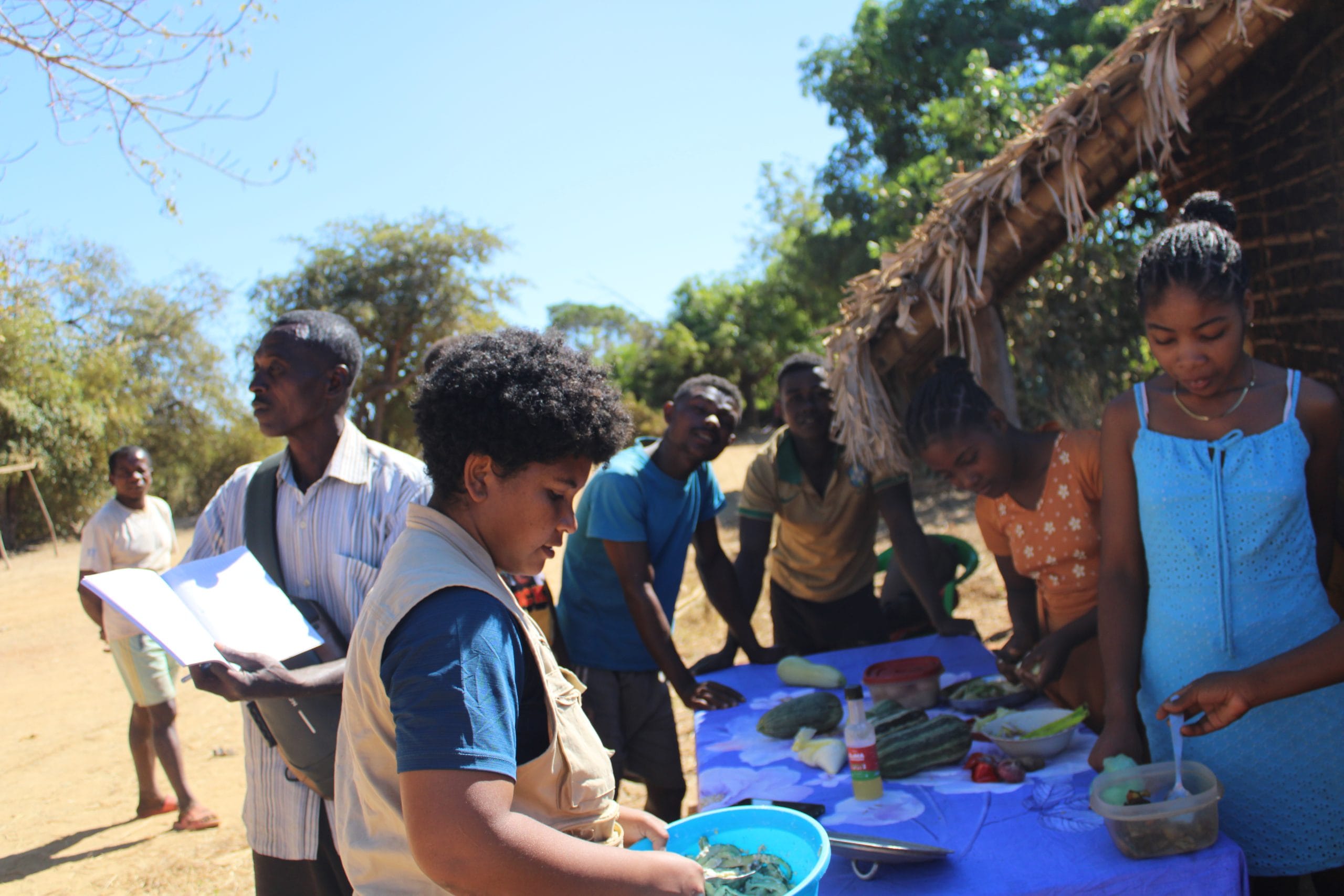 Group of people, including children, gathered around a table with bowls and food outdoors, near a thatched structure.