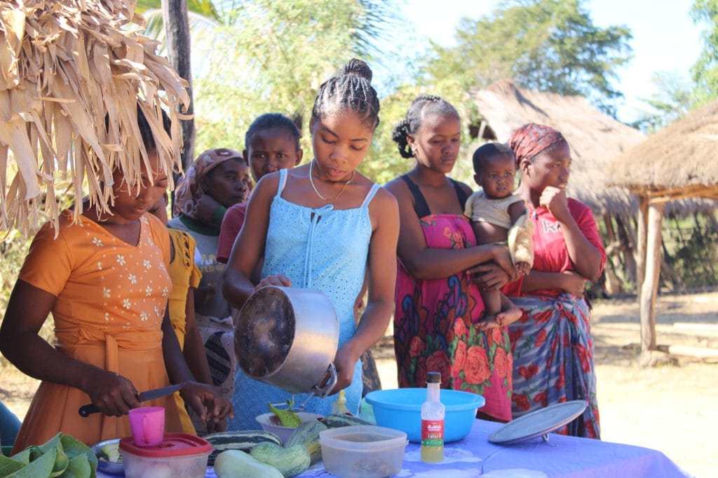 Group of women and children gathered around a table with bowls and vegetables for an outdoor meal in a rural village on a sunny day papering a meal prep scene