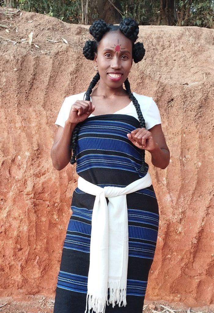Felice Anjarasoa Woman smiling at the camera, wearing a white top with a blue striped traditional wrap and white sash, standing before a dirt wall. Her hair is styled in large braided buns with two long braids framing her face, and a red forehead mark is visible.