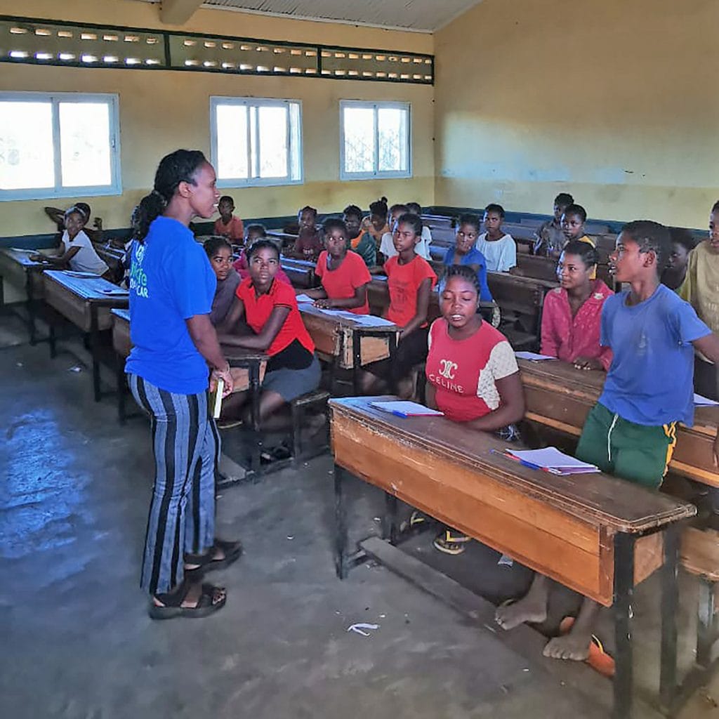Felice Anjarasoa Female teacher in a blue shirt speaks to a classroom of students seated at wooden desks in a yellow-walled room.
