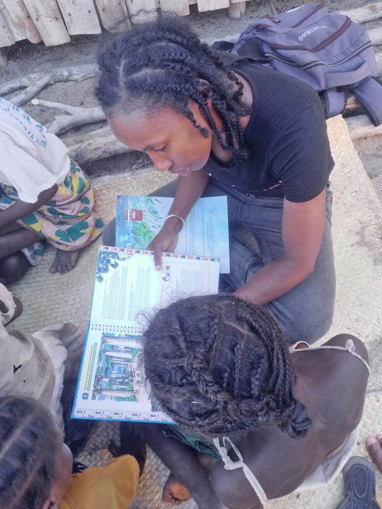 Felice Anjarasoa Young woman with braided hair reads a colorful workbook while others sit nearby on a woven mat outdoors.
