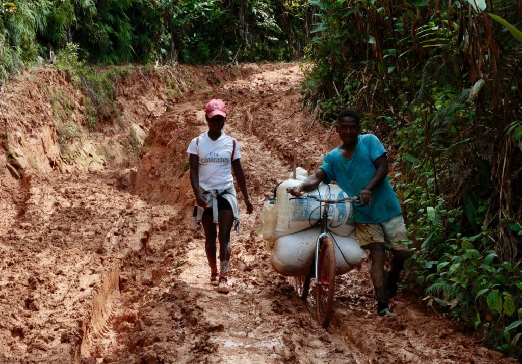 Two people push a bicycle overloaded with bags along a muddy, rutted jungle path.