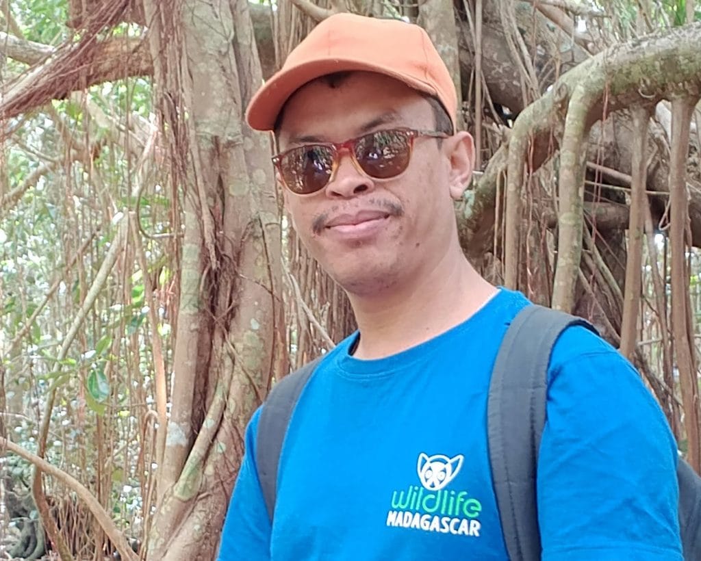 Man wearing orange cap, sunglasses, and a blue t-shirt with 'Wildlife Madagascar' logo, standing in a forest with vines and tree roots, carrying a backpack.